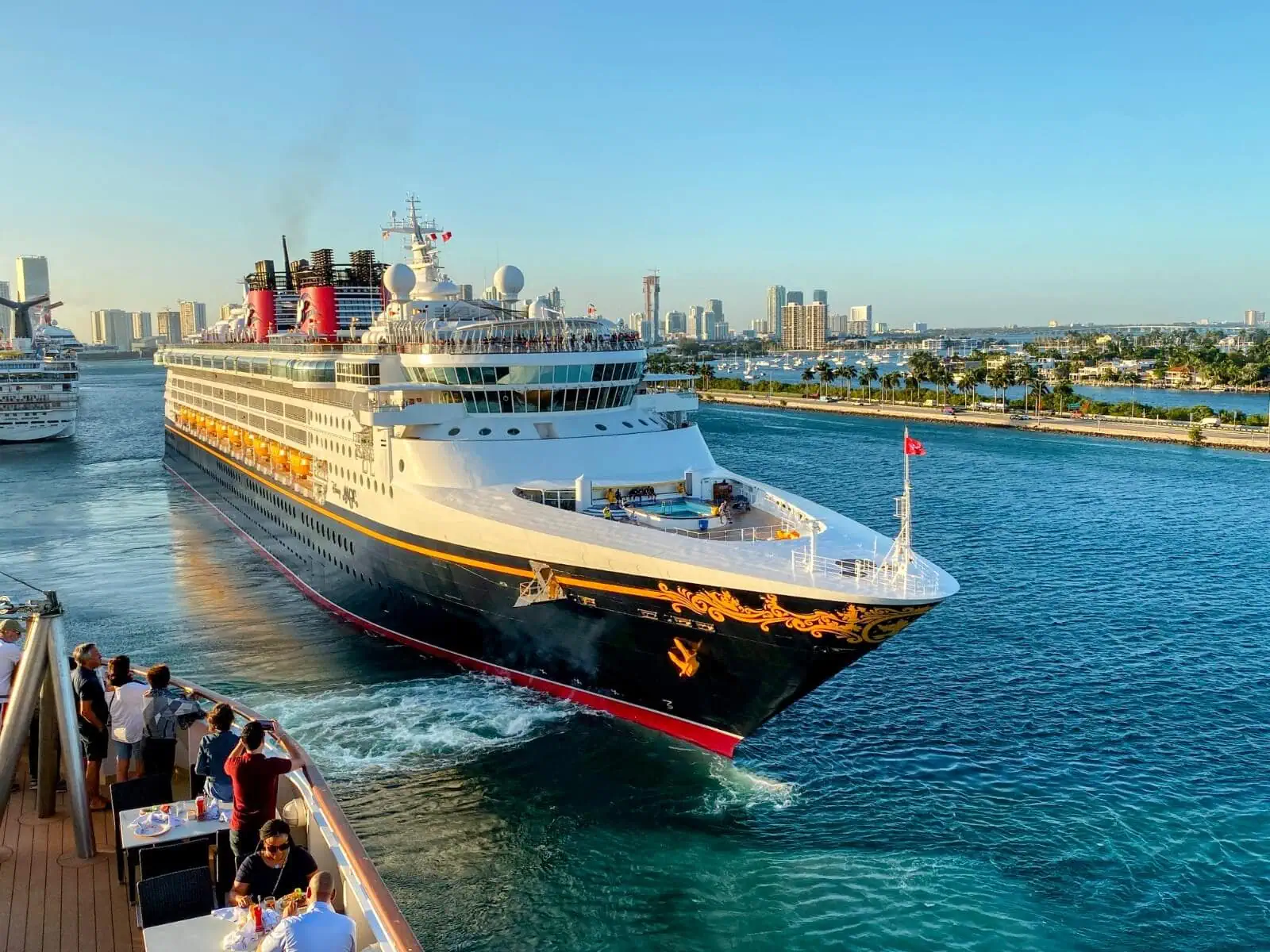 All aboard a large cruise ship sailing through a scenic waterway with a city skyline in the background. Passengers on board another Disney vessel watch and take photos, capturing the magical moment.