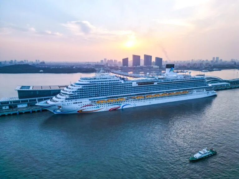 A large Chinese cruise ship docked at a port during sunset, with a city skyline in the background and a smaller boat nearby, created an air of mystery as it was revealed that a male passenger was missing.