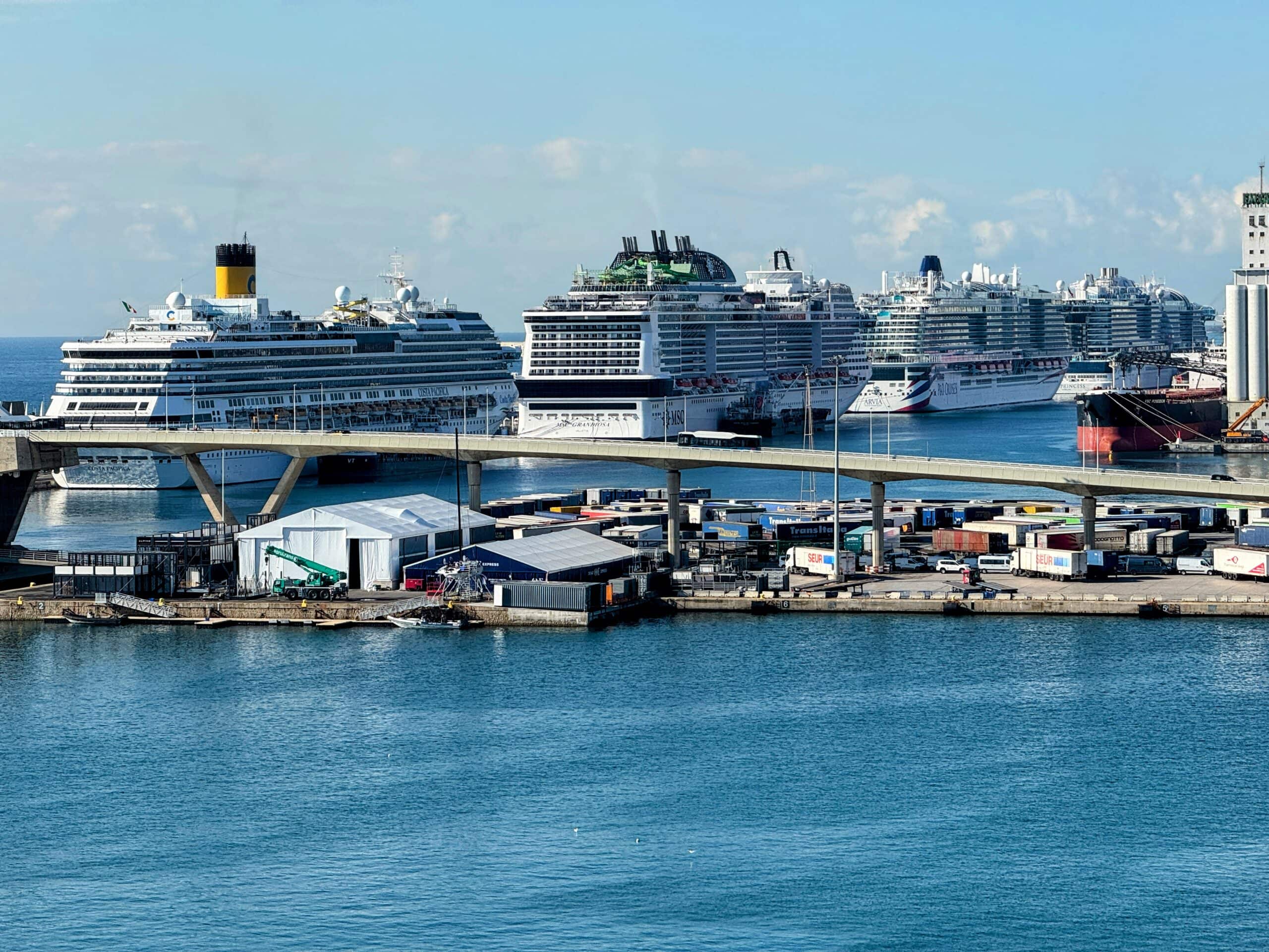 Several large cruise ships docked at the port of Barcelona, with an overpass bridge in the foreground and various buildings and shipping containers nearby.