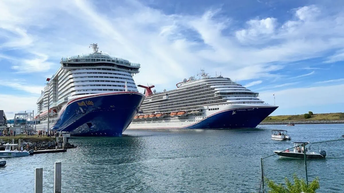 Two large cruise ships are docked at a port under a blue sky with light clouds. A small boat is on the water near the ships.