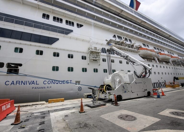 A large cruise ship called "Carnival Conquest" is docked at the cruise port for maintenance work, with equipment on the pier. Cones and barricades are placed around the area to ensure safety while showcasing its impressive shore power capabilities.