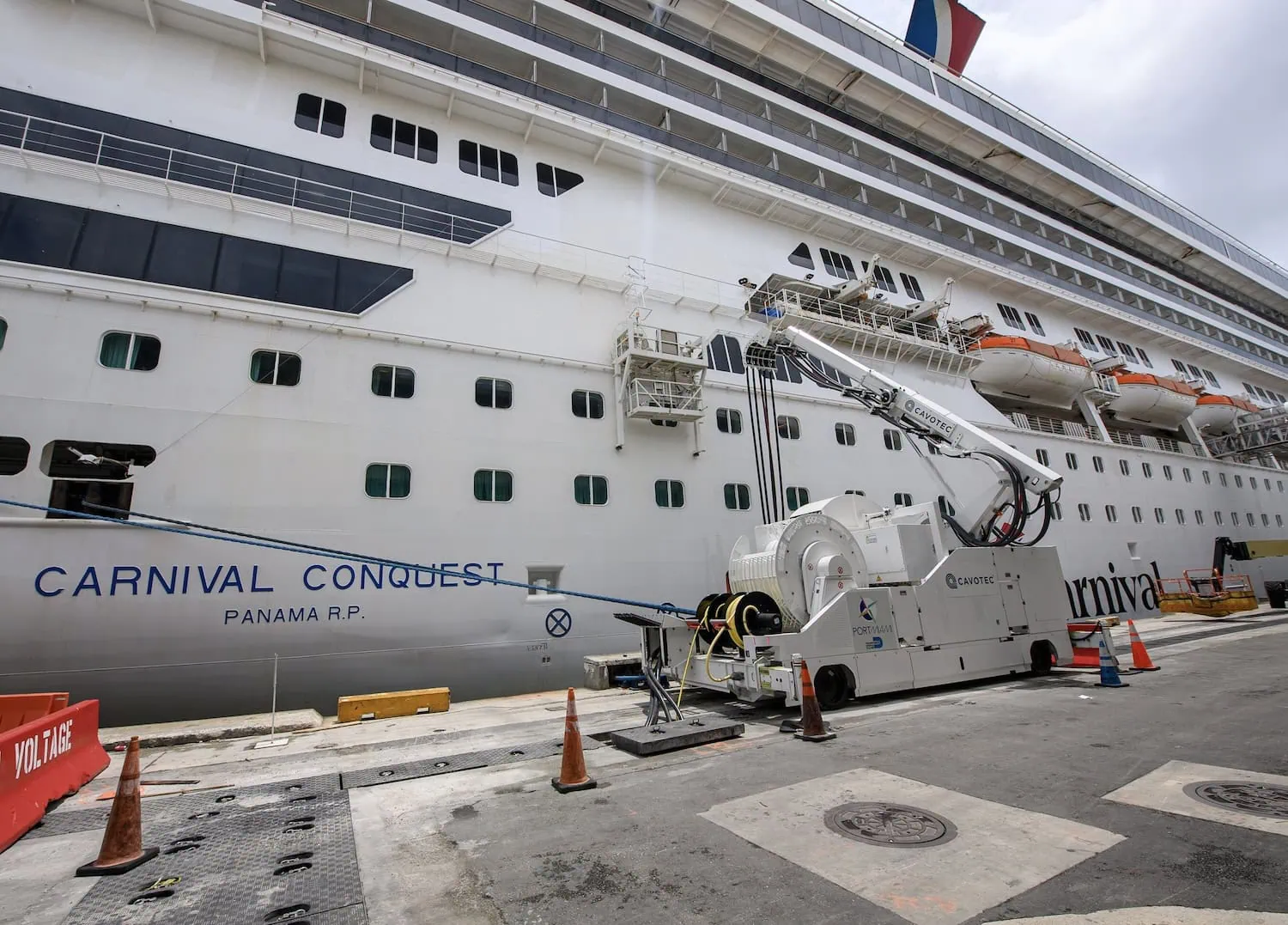 A large cruise ship called "Carnival Conquest" is docked at the cruise port for maintenance work, with equipment on the pier. Cones and barricades are placed around the area to ensure safety while showcasing its impressive shore power capabilities.