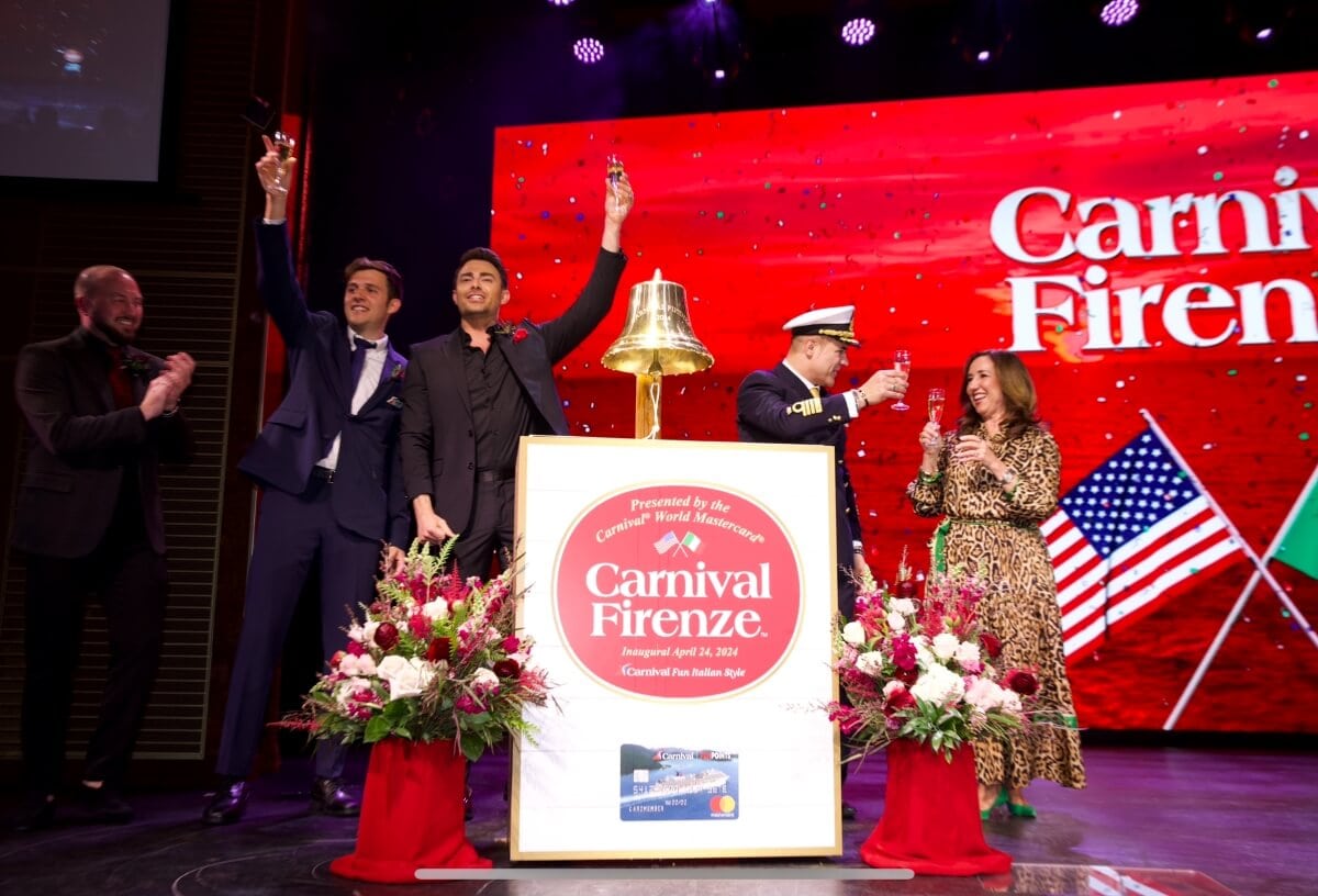 People celebrating at a ship's naming ceremony with a large bell and floral decorations on stage, under a sign that reads