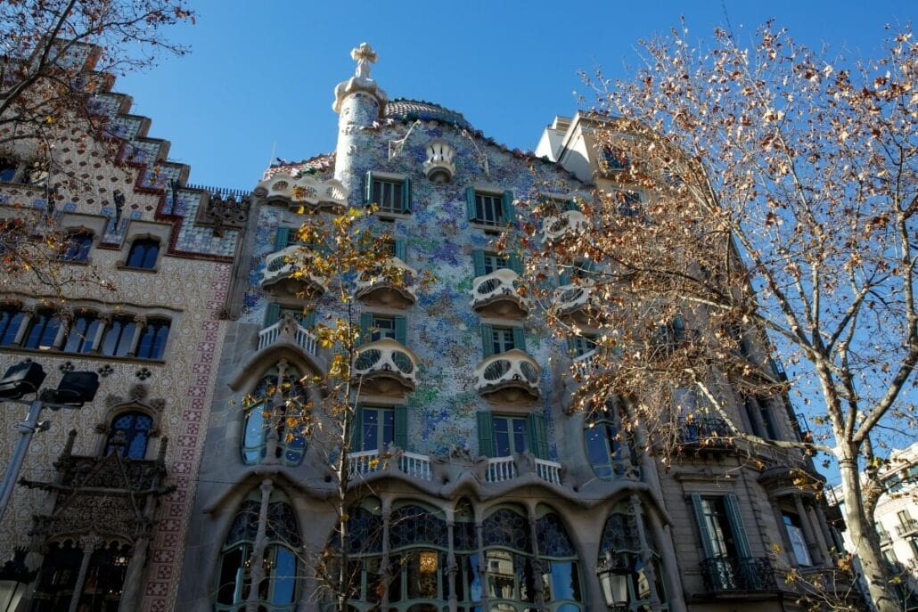 A multi-story building with an elaborate, colorful facade featuring organic shapes and windows, with bare trees in the foreground against a clear blue sky—one of the must-see sights for those exploring things to do in Barcelona.
