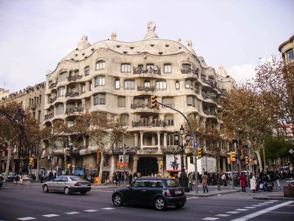A street view of Casa Milà in Barcelona, Spain, showcasing its unique undulating stone facade and wrought iron balconies, with cars and pedestrians at an intersection in the foreground—one of the must-see things to do for cruisers exploring the city's architectural wonders.