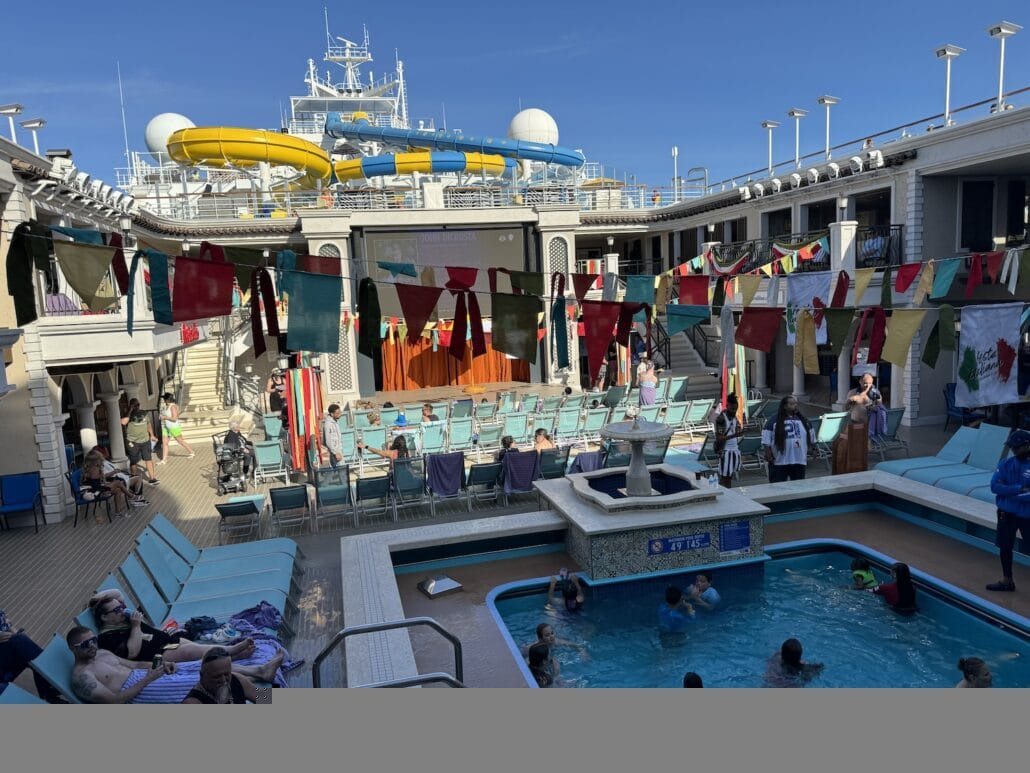 Cruise ship deck with passengers enjoying a pool, surrounded by colorful flags and deck chairs, with water slides visible in the background.