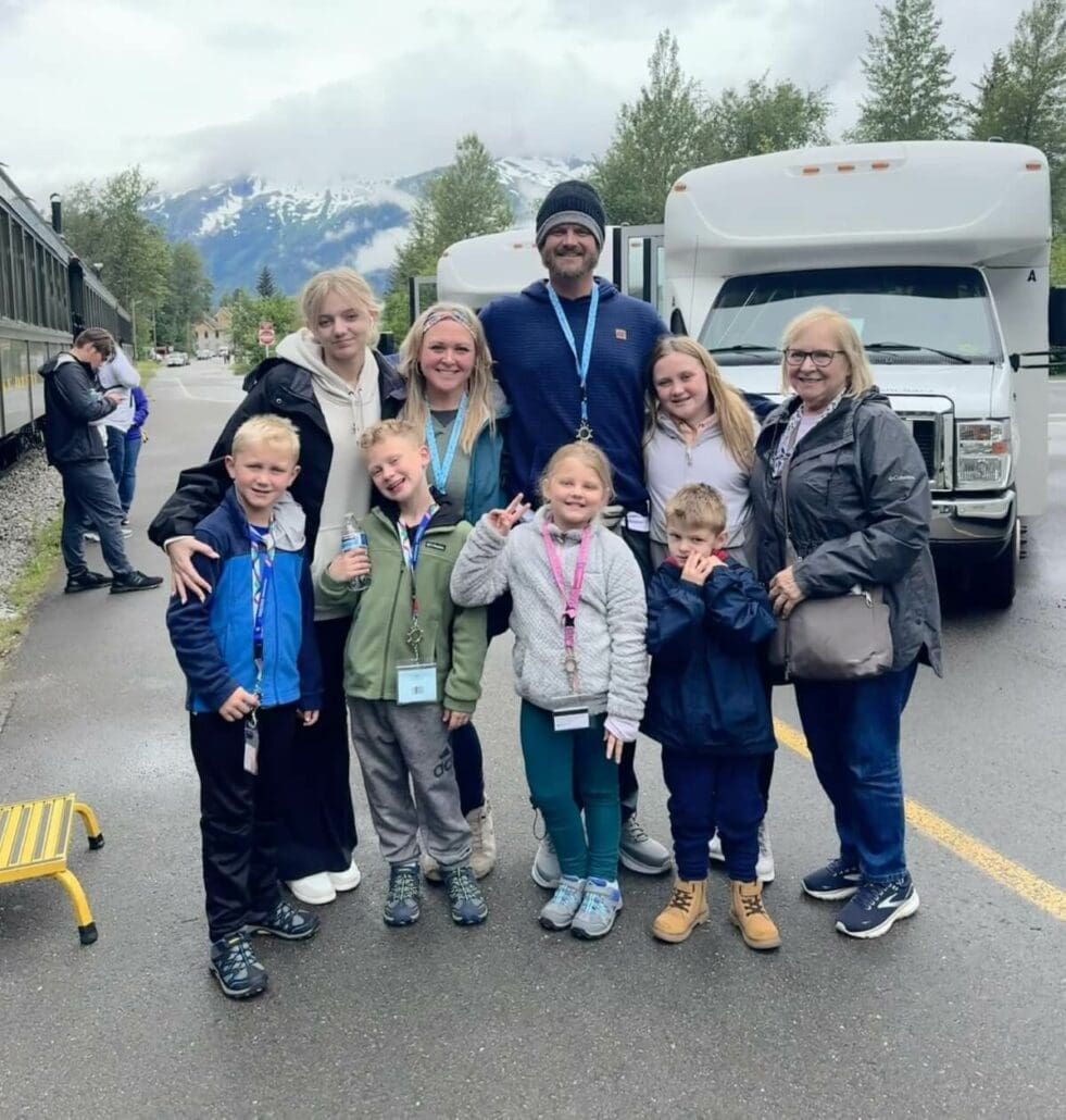 A family of 9, including adults and children, poses for a photo outdoors on a wet, overcast day with buses and a snow-capped mountain in the background.