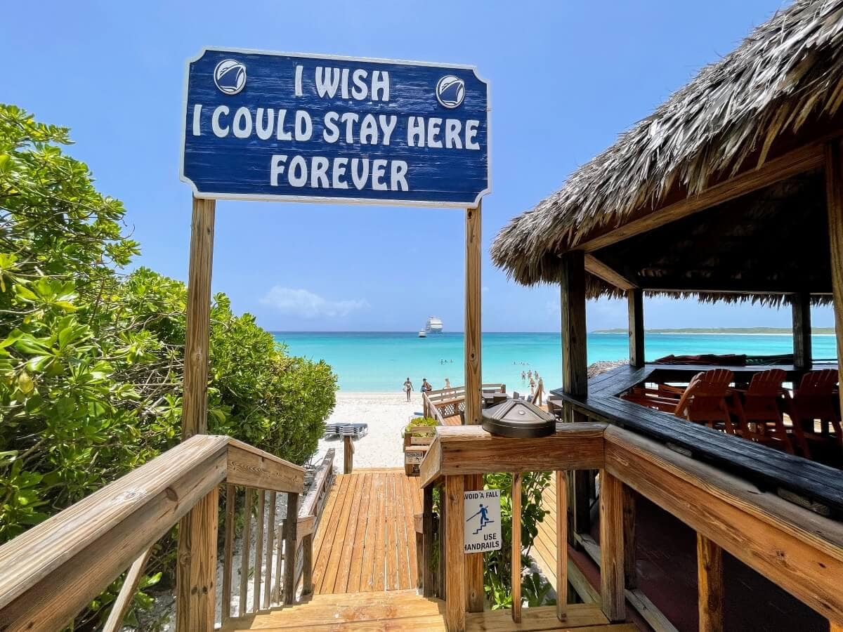 A pathway with wooden railings leads to a beach with turquoise water and a blue sky. A sign above reads