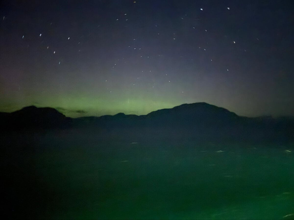 A night sky in Alaska shows faint stars above silhouetted mountains, with a hint of green aurora near the horizon.