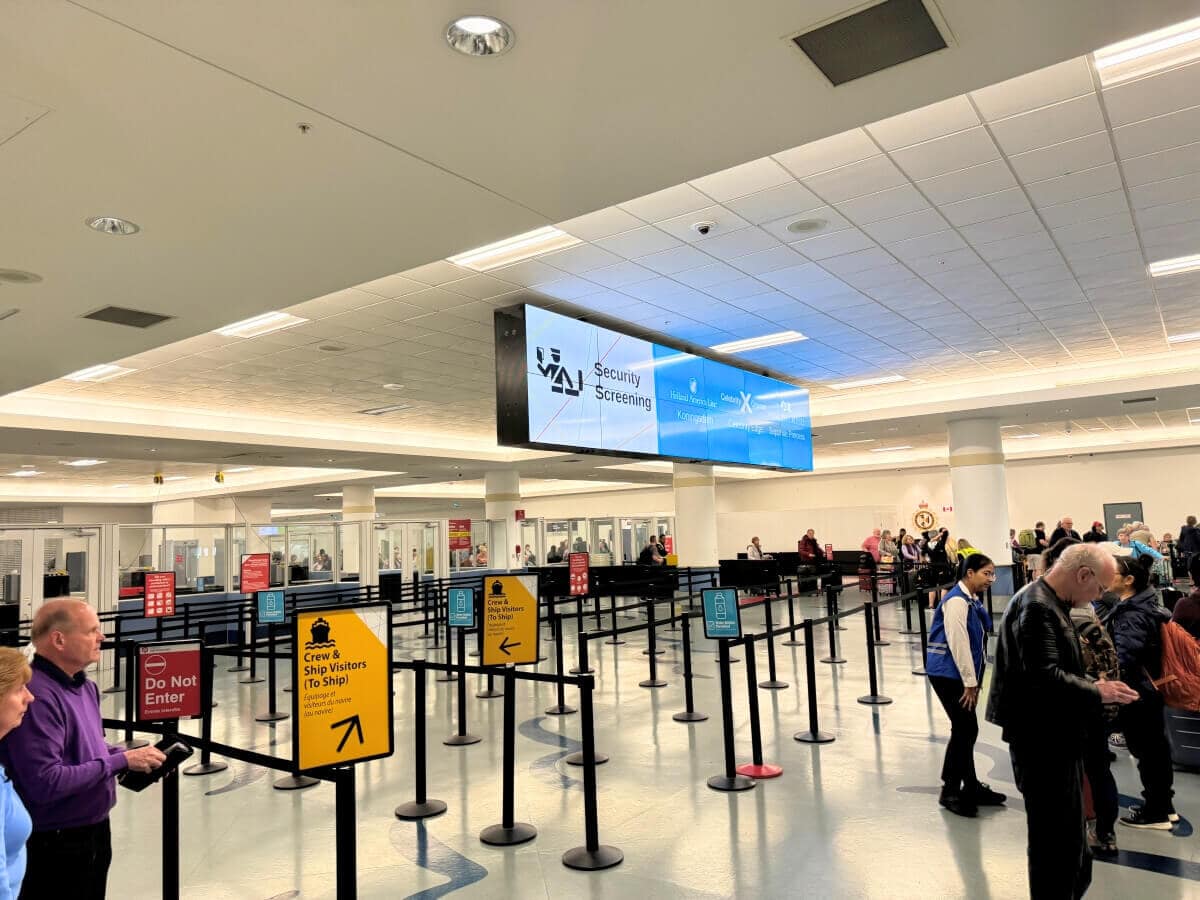 Airport security checkpoint with passengers lining up, guided by stanchions and informational signs. A large digital screen displays