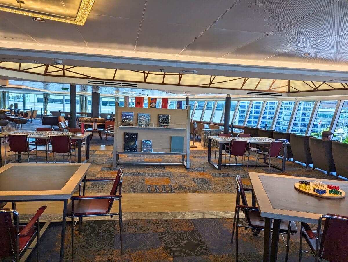 Interior of a modern library on the Koningsdam, featuring seating areas, shelves with books, and large windows overlooking city buildings.