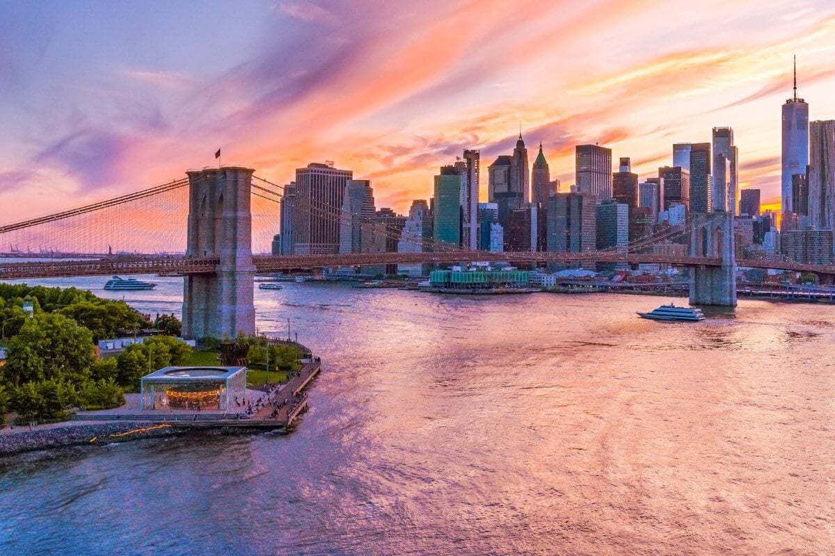 Aerial view of Brooklyn Bridge and Manhattan skyline at sunset, featuring colorful clouds and the New Virgin Ship, Brilliant Lady, reflecting on the water.