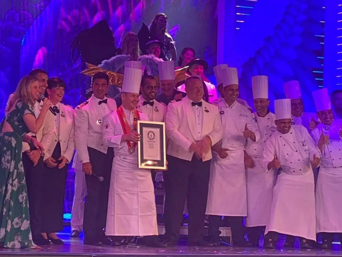 A group of chefs and individuals in white attire holds a framed certificate on a brightly lit stage, with some clapping and smiling, celebrating their achievement at the Princess Cruises' Giant Pizza Party that set a new Guinness World Record.