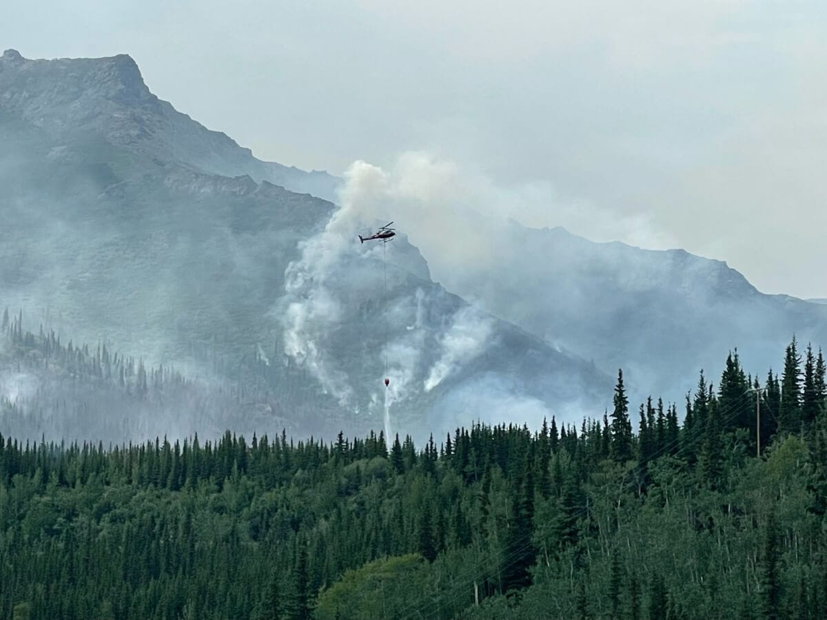 A helicopter carries water to combat a wildfire in a mountainous forest area, with smoke billowing from burning trees below, revealing the urgent nature of the operation.