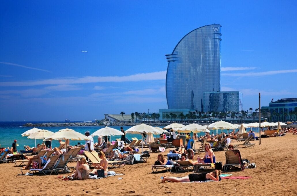 People relaxing on a sandy beach with umbrellas and lounging chairs. In the background, a large, modern hotel rises against a clear blue sky—a perfect spot to unwind after enjoying cruise excursions or other Barcelona activities.