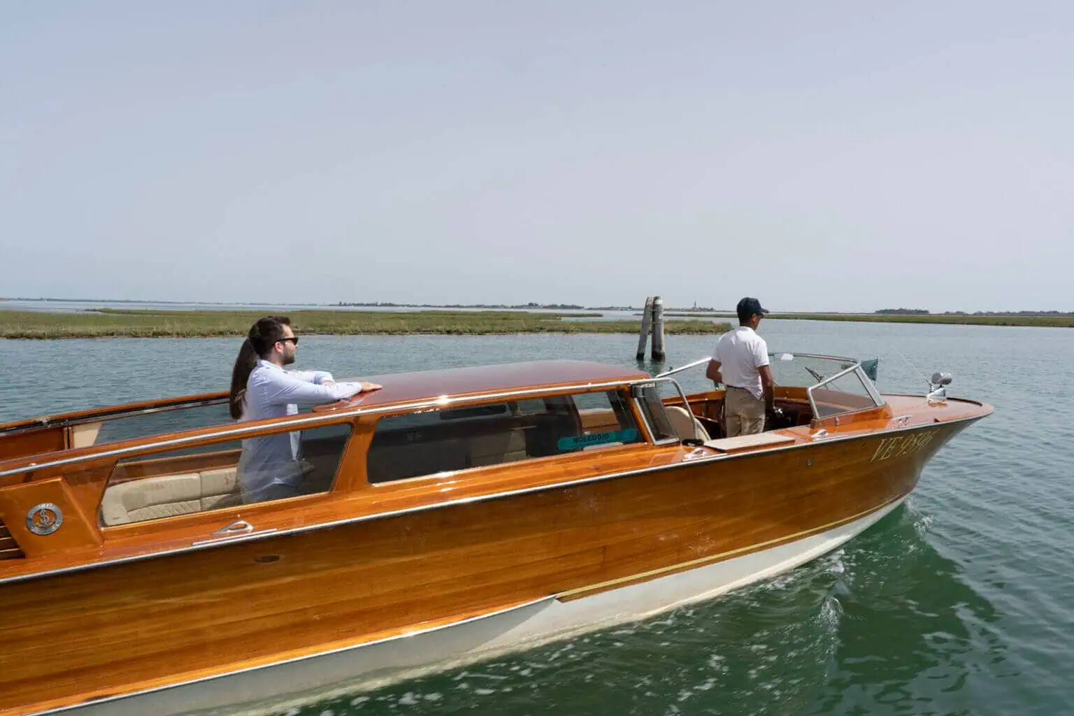 Two people ride on a wooden motorboat in a calm body of water near a marshy coastline. One stands at the helm, while the other rests by the railing, dreaming of their next European cruise.