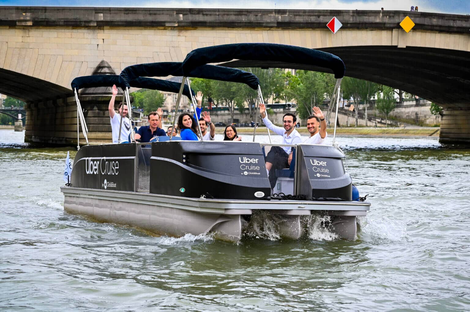 A group of people wave from a boat with a canopy labeled